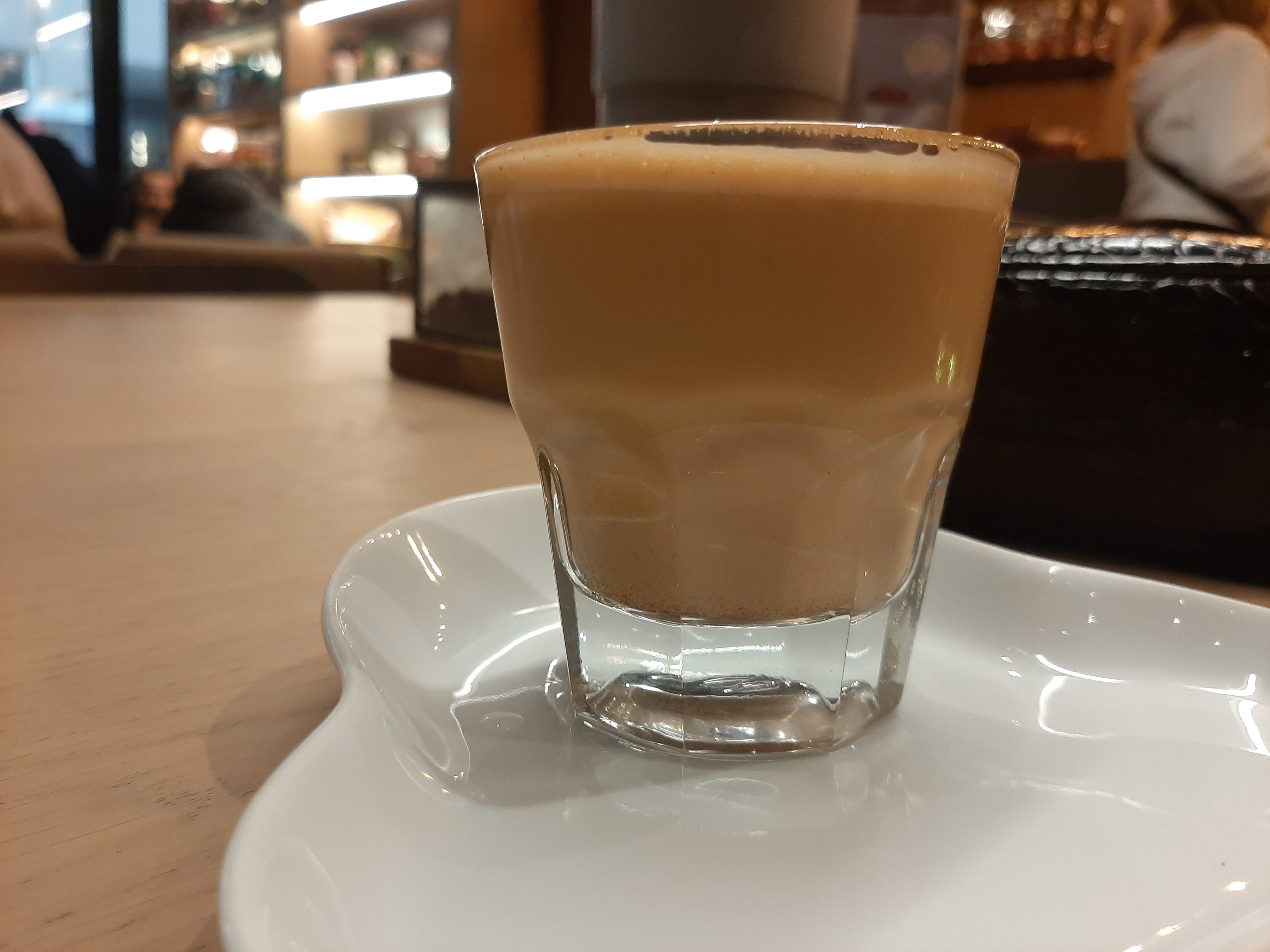 A glass of cortado coffee on a white saucer, placed on a wooden table in a cosy café. Blurred shelves and people are in the background
