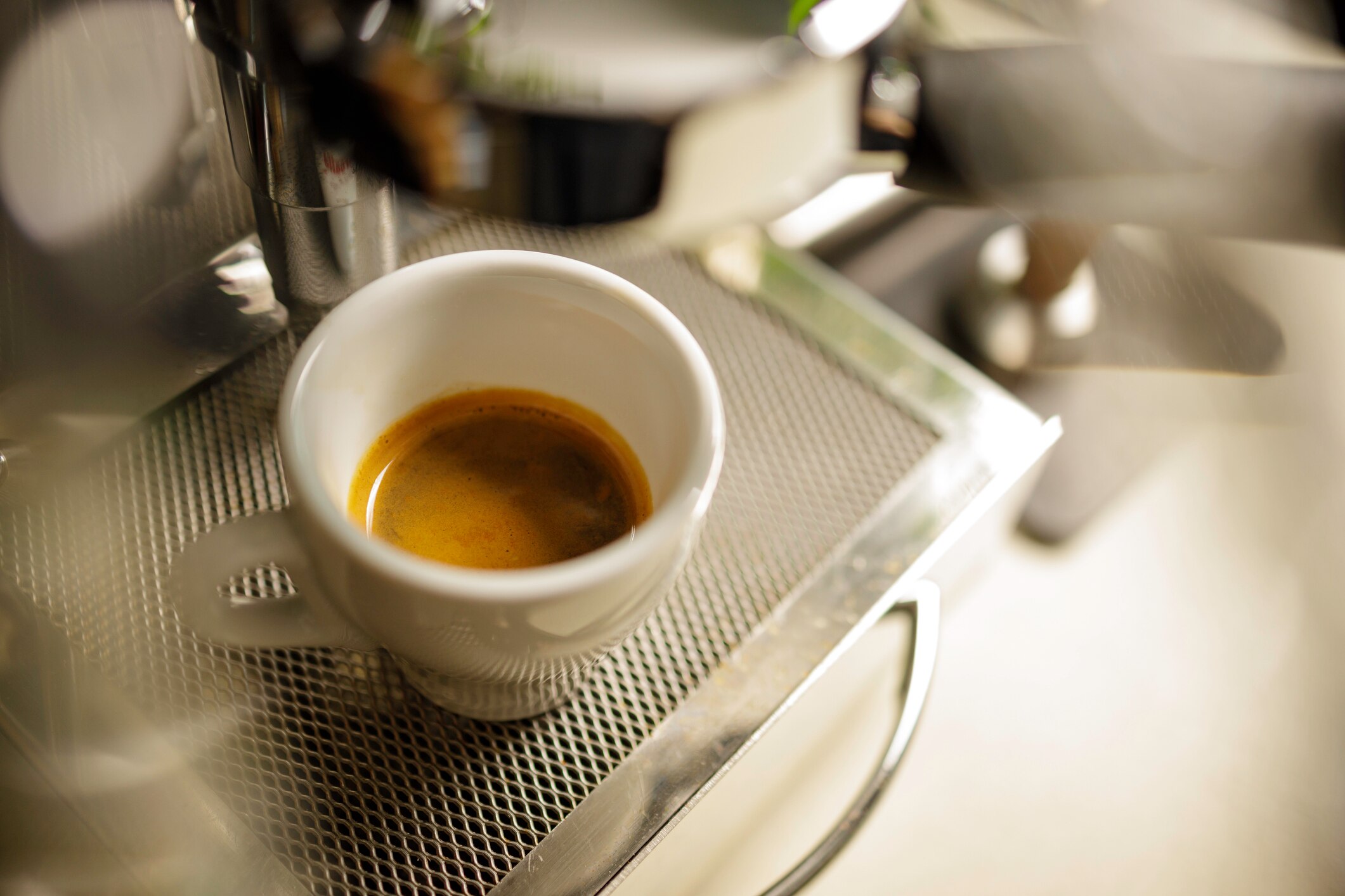 A freshly brewed espresso shot in a white cup, placed on a metal tray of an espresso machine.