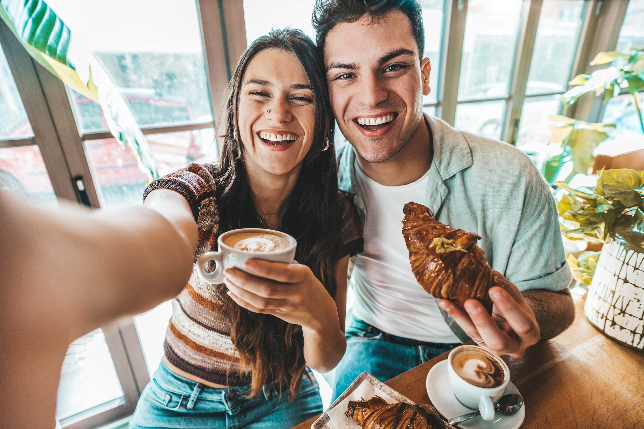 A woman on the left holding a mug of macchiato, smiling next to a man on the right holding a croissant